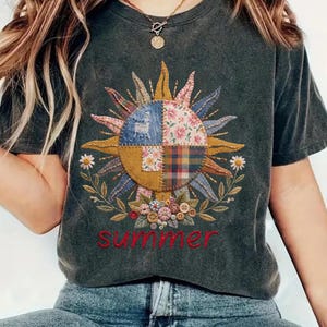 以下が含まれることがあります： カラフルな太陽のデザインが特徴の濃いグレーのTシャツ。花柄とパッチワークの要素が含まれています。「summer」の文字が太陽の下に赤いスクリプトで書かれています。このシャツは半袖で、クルーネックです。