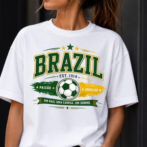 May include: A white t-shirt featuring a graphic design with the word BRAZIL in green and yellow block letters. Below, a soccer ball sits between green and yellow brush strokes with the text PAIX&Atilde;O, ORGULHO, EST. 1914, and UM PAIS. UMA CAMISA. UM SONHO.
