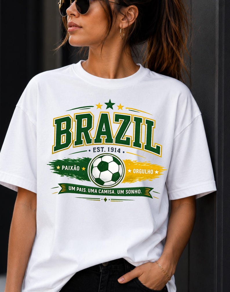 May include: A white t-shirt featuring a graphic design with the word BRAZIL in green and yellow block letters. Below, a soccer ball sits between green and yellow brush strokes with the text PAIX&Atilde;O, ORGULHO, EST. 1914, and UM PAIS. UMA CAMISA. UM SONHO.