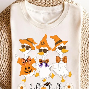 Op de afbeelding: Crèmekleurig T-shirt met drie cartoon ganzen in Halloween kostuums. De ganzen dragen oranje hoeden en zonnebrillen, met outfits met pompoen-, heksen- en spookthema's. De tekst "hello fall" staat in een scriptletter onder de ganzen.