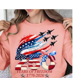 Puede incluir: Una sudadera de color coral con un diseño patriótico. El gráfico incluye aviones a reacción, una bandera estadounidense estilizada y el número "250" en rojo, blanco y azul. El texto dice "YEARS OF FREEDOM 1776-2026".