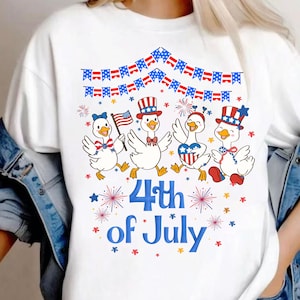 Può includere: T-shirt bianca con un design del 4 luglio. La grafica mostra quattro paperi dei cartoni animati in abiti patriottici, che tengono bandiere e fuochi d'artificio, con la scritta "4th of July" in blu.