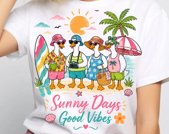 May include: White t-shirt featuring a cartoon illustration of four ducks on a beach, each wearing sunglasses and beach attire. The shirt has the text "Sunny Days Good Vibes" in teal and pink.
