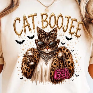 May include: A cream-colored t-shirt featuring a graphic of a cat wearing leopard print sunglasses, a matching bow, and a faux fur coat. The cat holds a coffee cup and a pink leopard print purse. The text CAT BOOJEE appears in gold above.