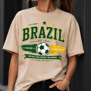 May include: A beige short-sleeve t-shirt featuring a graphic with the text BRAZIL, EST. 1914, PAIX&Atilde;O, ORGULHO, and UM PAIS. UMA CAMISA. UM SONHO. The design includes a soccer ball, green and yellow brush strokes, and stars in a collegiate font.