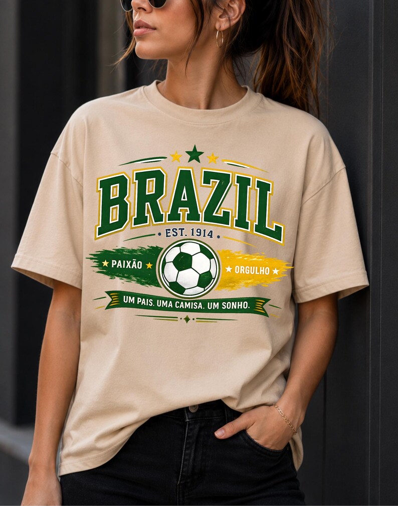 May include: A beige short-sleeve t-shirt featuring a graphic with the text BRAZIL, EST. 1914, PAIX&Atilde;O, ORGULHO, and UM PAIS. UMA CAMISA. UM SONHO. The design includes a soccer ball, green and yellow brush strokes, and stars in a collegiate font.