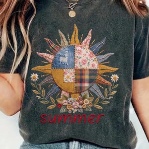May include: A charcoal gray t-shirt featuring a central sun graphic composed of patchwork fabric squares in denim, floral, and plaid patterns. The sun is surrounded by embroidered leaves, daisies, and buttons, with the word summer in red text below.