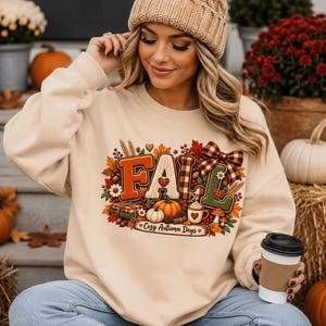 Op de afbeelding: Een beige sweater met ronde hals en een grafische print die het woord FALL spelt met herfstthema-elementen zoals pompoenen, bladeren, een geruite strik en een mok cacao. Onder de afbeelding staat de tekst Cozy Autumn Days in een schrijfletter.