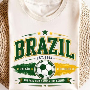 May include: A cream-colored t-shirt featuring a graphic with the word BRAZIL in green and yellow block letters. Below it, the text EST. 1914, a soccer ball, and the words PAIX&Atilde;O, ORGULHO, and UM PAIS. UMA CAMISA. UM SONHO. are printed in green and yellow.