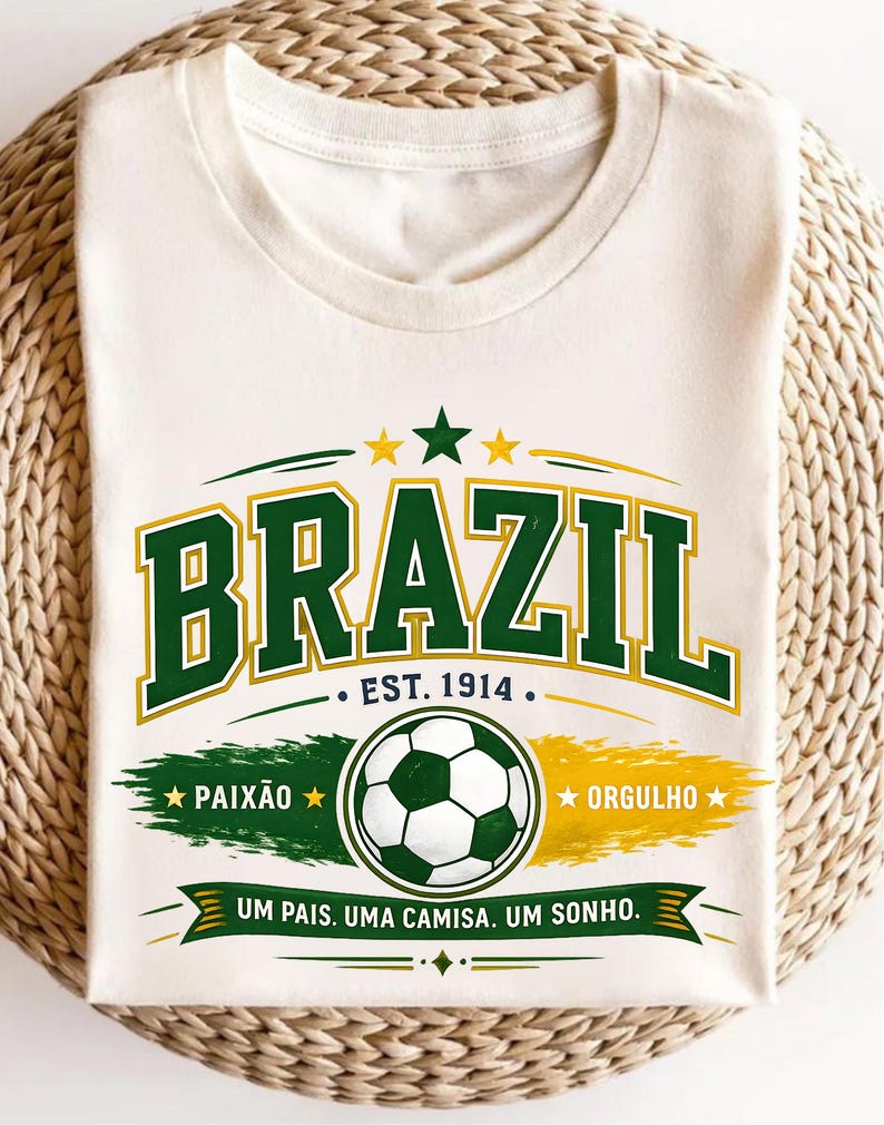 May include: A cream-colored t-shirt featuring a graphic with the word BRAZIL in green and yellow block letters. Below it, the text EST. 1914, a soccer ball, and the words PAIX&Atilde;O, ORGULHO, and UM PAIS. UMA CAMISA. UM SONHO. are printed in green and yellow.