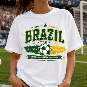 May include: A white short-sleeved t-shirt featuring a graphic print with the word BRAZIL in green and yellow, a soccer ball, the text EST. 1914, PAIX&Atilde;O, ORGULHO, and the phrase UM PAIS. UMA CAMISA. UM SONHO. in green, yellow, and white colors.