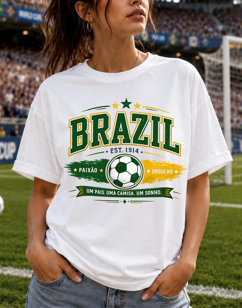 May include: A white short-sleeved t-shirt featuring a graphic print with the word BRAZIL in green and yellow, a soccer ball, the text EST. 1914, PAIX&Atilde;O, ORGULHO, and the phrase UM PAIS. UMA CAMISA. UM SONHO. in green, yellow, and white colors.