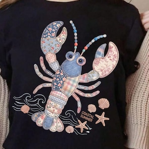 May include: Black t-shirt featuring a patchwork lobster design with floral and plaid patterns. The lobster has blue, pink, and white accents, with the words "Coastal Summer" and seashell and starfish graphics.