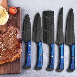 May include: A set of six black chef knives with textured blades and blue handles. The handles have gold-colored rivets. The knives are displayed on a wooden cutting board next to a steak, tomato, and onion.