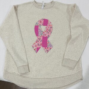 May include: A light beige sweatshirt with a pink ribbon made of patchwork fabric. The ribbon is a symbol of breast cancer awareness.