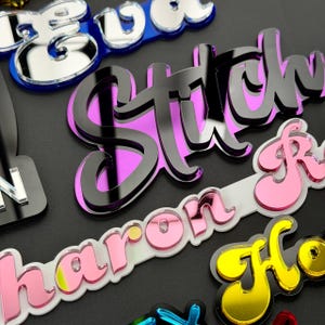 May include: A collection of layered acrylic name tags featuring various colors including black, pink, yellow, and blue. Each tag displays custom text in a cursive script font, with a reflective mirrored finish on the top layer and a solid base.