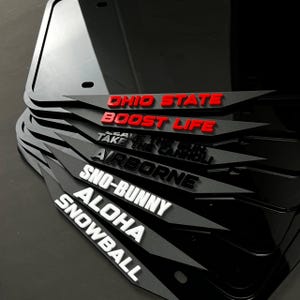 以下が含まれることがあります： 赤と白の文字デザインが施された黒いナンバープレートフレームのスタック。「OHIO STATE」、「BOOST LIFE」、「AIRBORNE」、「SNO-BUNNY」、「ALOHA」、「SNOWBALL」などのフレーズが含まれています。