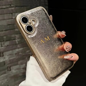 May include: Gold-colored phone case with a textured surface and the initials "A.M." in gold lettering. The case has a gold frame around the camera lenses. The phone case is held in a hand.