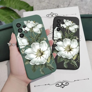 Op de afbeelding: Twee smartphonehoesjes met een botanische illustratie van witte magnolia bloemen met groene bladeren. Het ene hoesje heeft een gedempte saliegroene achtergrond, terwijl het andere een zwarte achtergrond heeft. Beide hoesjes hebben een camera-uitsparing.