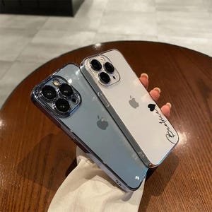 May include: Two clear phone cases for an iPhone. The case on the left is a light blue colour with a black camera lens. The case on the right is clear with a black heart and the text "Forever" written on it.