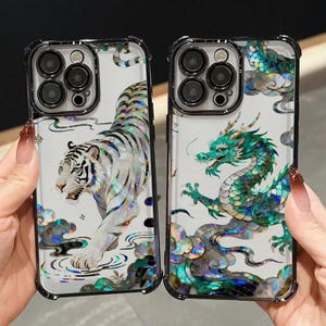 Può includere: Due custodie trasparenti per telefono con bordi neri. Una presenta un design di tigre bianca, l'altra un drago verde. Entrambi i design hanno accenti iridescenti e sono su uno sfondo nuvoloso.