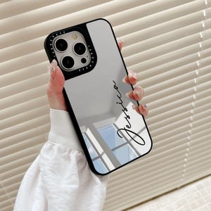 Può includere: Una custodia per telefono a specchio nero con una scritta bianca che dice "Jessica". La custodia ha un bordo nero attorno agli obiettivi della fotocamera e un cerchio nero attorno agli obiettivi con le parole "Pretty Eye" scritte in bianco.