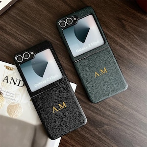 Può includere: Due custodie per telefono nero e verde con una finestra trasparente sul davanti. Le custodie sono progettate per il telefono Galaxy Z Flip6. Le custodie hanno le iniziali "A.M." in rilievo sul retro.