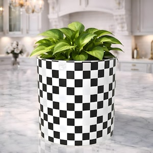 May include: A round plant pot with a black and white chequered design. The pot contains green leafy plants. The pot is on a marble countertop in a kitchen setting.