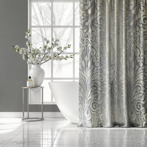 May include: A bathroom scene featuring a white bathtub, a silver side table with a white vase of flowers, and a patterned gray and white curtain. The window behind the tub offers a view of a tree.