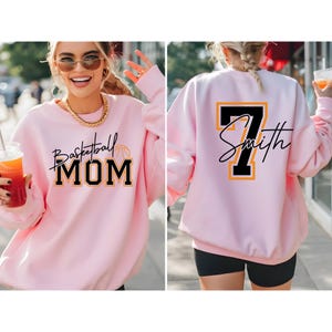 May include: A light pink sweatshirt with the words "Basketball MOM" in black script and block letters, with a basketball graphic. The back of the sweatshirt has the number 7 and the name "Smith" in black and yellow.