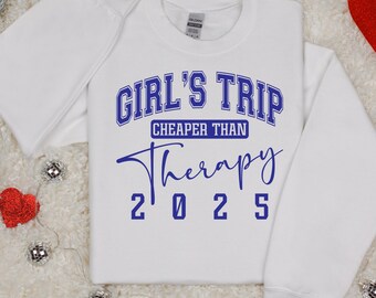 May include: White sweatshirt with the text "GIRL'S TRIP CHEAPER THAN Therapy 2025" in blue. The sweatshirt is folded and placed on a white, textured surface, with decorative silver and red accents.