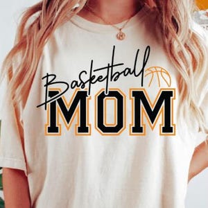 May include: Cream-colored t-shirt with the words "Basketball MOM" in black and orange lettering. A basketball graphic is above the word "MOM". The shirt is a casual style, suitable for sports fans.