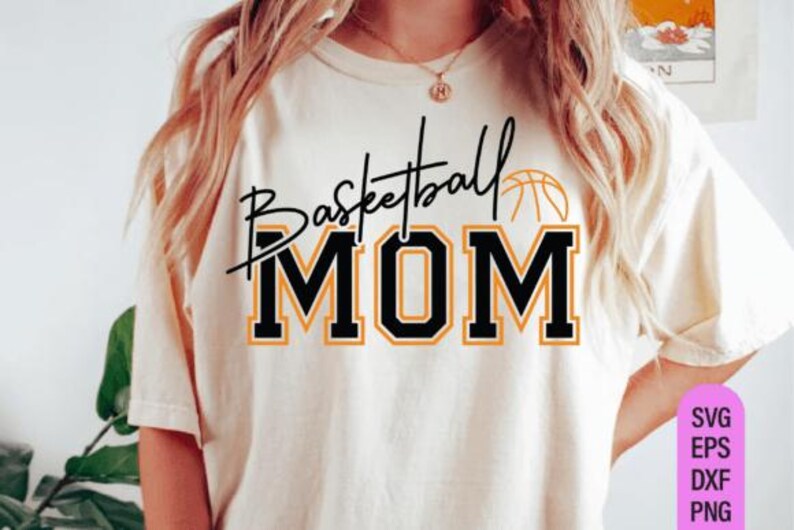 May include: Cream-colored t-shirt with the words "Basketball MOM" in black and orange lettering. A basketball graphic is above the word "MOM". The shirt is a casual style, suitable for sports fans.