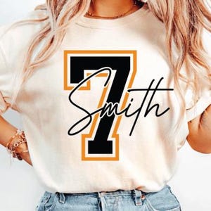 May include: Cream-colored t-shirt featuring a large black number 7 with an orange outline. The name "Smith" is written in a cursive font across the number. The shirt design is likely for sports or team-related apparel.