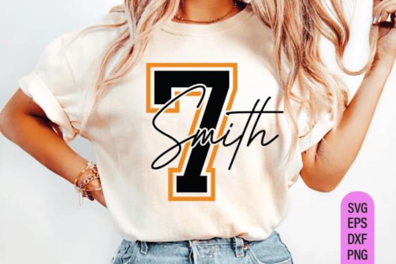 May include: Cream-colored t-shirt featuring a large black number 7 with an orange outline. The name "Smith" is written in a cursive font across the number. The shirt design is likely for sports or team-related apparel.