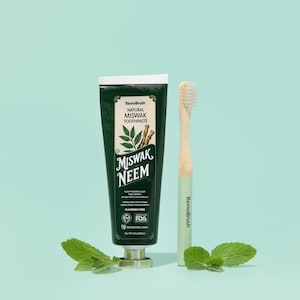 Puede incluir: Un tubo verde oscuro de pasta dental Remobrush Natural Miswak Neem y un cepillo de dientes a juego con un mango verde claro. El tubo tiene una tapa blanca y una base plateada. Hojas de menta fresca se colocan alrededor de los productos.