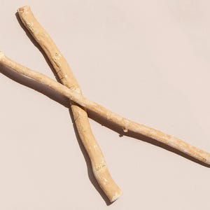 May include: Two light brown miswak sticks, also known as siwak, are crossed against a light pink background. One stick has a frayed end, and the other two are smooth. These natural toothbrushes are used for oral hygiene.