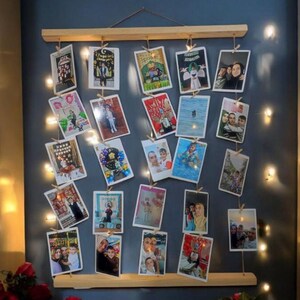 Peut inclure: Un présentoir photo en bois avec plusieurs photos suspendues à de la ficelle. L'ensemble est monté sur un mur bleu foncé et éclairé par de petites guirlandes lumineuses. Les photos sont de tailles variées et présentent des personnes et des événements.