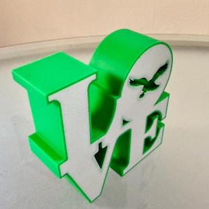 Könnte beinhalten: Grün-weißes 3D-gedrucktes "Love"-Schild mit einer Silhouette eines Vogels im Flug.
