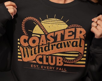 Peut inclure: Sweat-shirt noir avec un motif rétro orange et jaune. Le motif comprend le texte "Coaster Withdrawal Club", un manège de montagnes russes, un soleil et la phrase "Est. Every Fall."