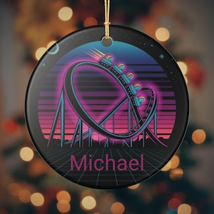 May include: A black ceramic Christmas ornament with a retro roller coaster design. The coaster is depicted in shades of blue, pink, and purple against a gradient background. The name "Michael" is written in pink. A gold string is attached for hanging.