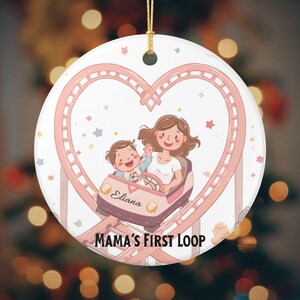 May include: A round ceramic Christmas ornament featuring a cartoon illustration of a mother and child riding a roller coaster. The roller coaster track forms a heart shape. The ornament has the text "Eliana" and "Mama's First Loop".