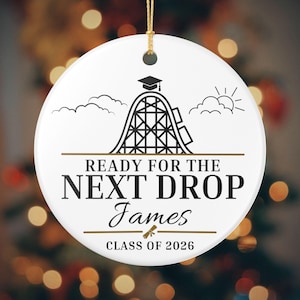 May include: White ceramic ornament with a gold string for hanging. The ornament features a black roller coaster graphic, clouds, and the text "READY FOR THE NEXT DROP James CLASS OF 2026". A graduation cap and diploma are also included.