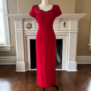 May include: A striking red, full-length dress with short sleeves and an ornate neckline. The dress is presented on a black mannequin, highlighting its elegant design. The background includes a fireplace and wooden flooring.
