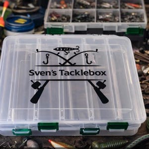 Könnte beinhalten: Eine transparente Angelkiste mit grünen Verschlüssen, mit dem schwarzen Schriftzug "Sven's Tacklebox". Die Kiste ist von Angelausrüstung umgeben, darunter Köder, Rollen und Schwimmer. Sie zeigt eine Grafik mit gekreuzten Angelruten und Haken.