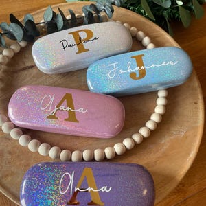 Könnte beinhalten: Vier personalisierte Brillenetuis in schillernden Farben: Weiß, Blau, Rosa und Lila. Jedes Etui hat einen goldenen Anfangsbuchstaben und Namen in Schreibschrift. Die Etuis sind auf einem Holztablett mit Perlenrand und Grün angeordnet.