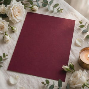Op de afbeelding: Een rechthoekig diepbordeauxrood kartonnen vel gecentreerd op een witte stoffen ondergrond, omringd door witte rozen, groene eucalyptus takjes en een kleine brandende kaars in een metalen gouden houder.