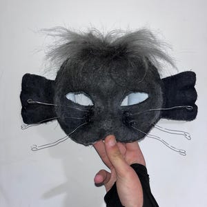 May include: A handmade black cat mask with gray fur and wire whiskers. The mask has large, open eye holes and is held up by a hand. The mask is made of a soft, felt-like material.