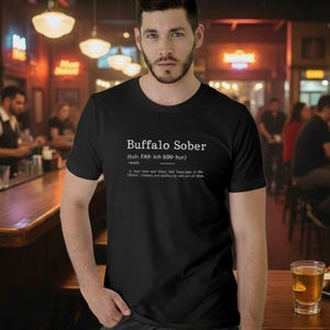 Può includere: T-shirt nero con la scritta bianca "Buffalo Sober". La maglietta ha una definizione sotto il titolo. L'immagine è scattata in un bar, con un uomo che indossa la maglietta in primo piano.