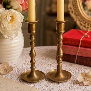 May include: Two matching brass candlesticks with a polished finish and a tiered, slender stem design. Each candlestick holds a single cream-colored taper candle. The brass metal has a warm, golden tone with a smooth, reflective surface texture.